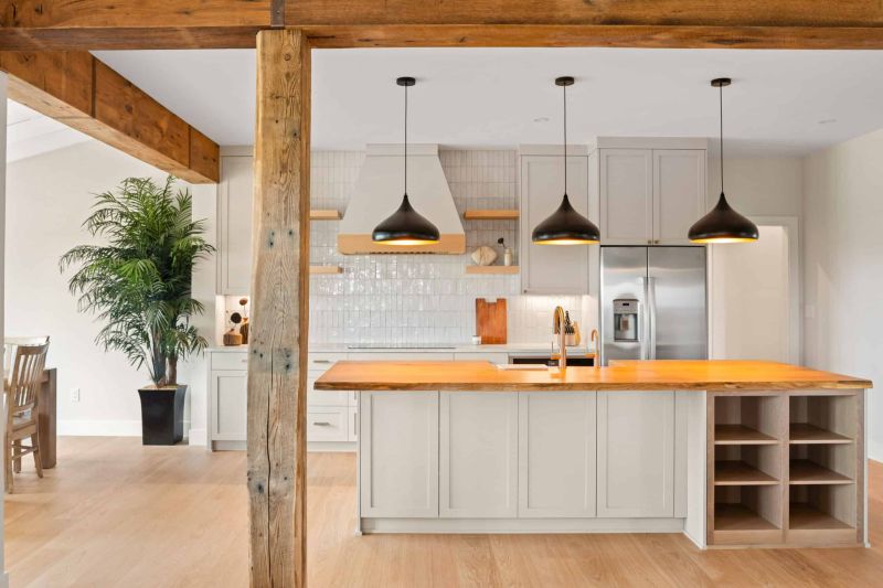 kitchen with shiny white tile walls off-white cabinets and an island with three hanging lights as seen framed between thick wood house beams