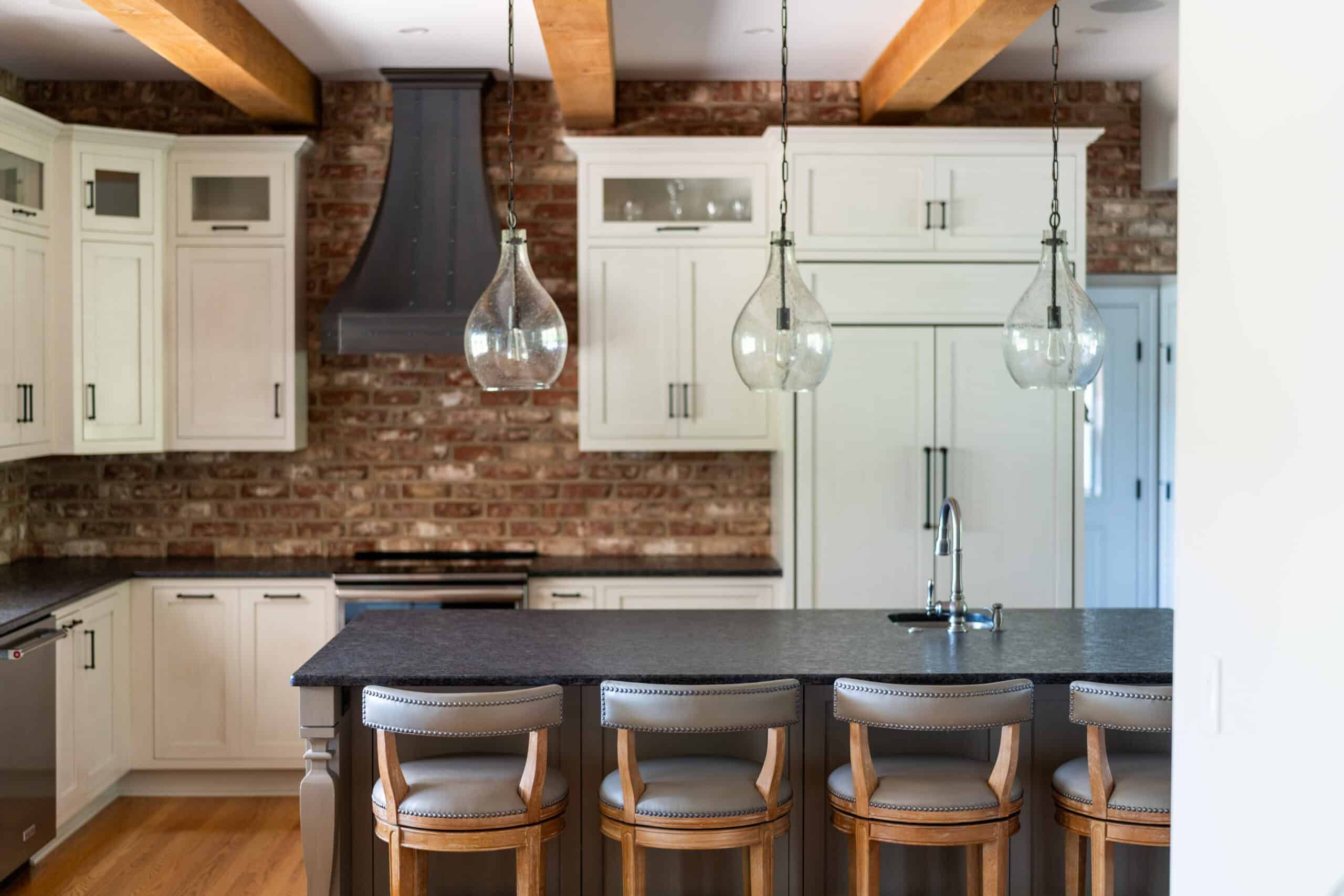 a large kitchen island with four wooden chairs with grey leather seats, three transparent glass lights hang from above, the kitchen has white cabinets and natural brick walls. A black granite stove hood and three thick wood ceiling beams are being showcased