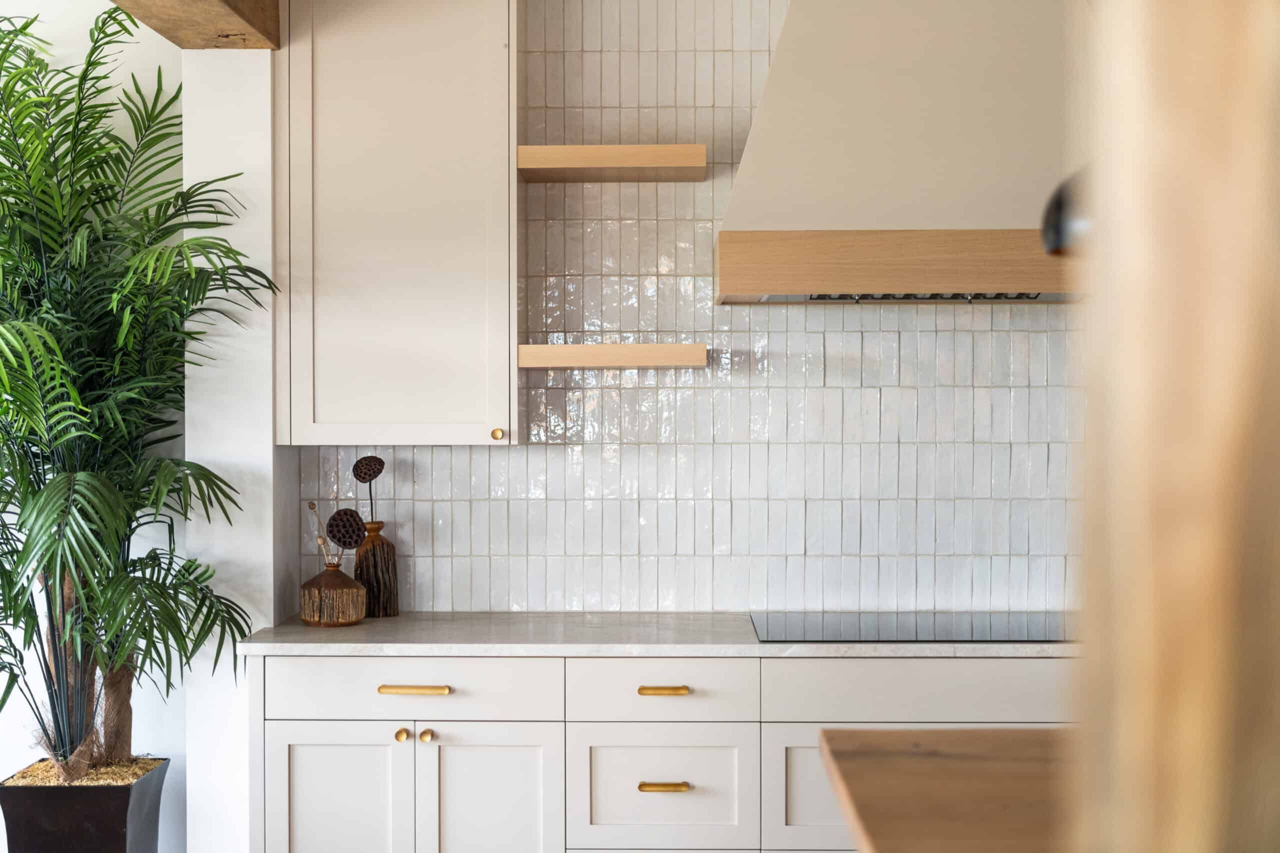 off-white kitchen cabinets with light brown shelves attached to a shiny white, vertical tile wall. A tall green floor plant is to the left