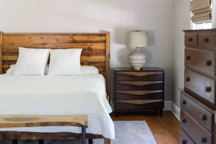 tight crop of a bed, with white coverings and light wood backboard, a dark wood nightstand with lamp, and a dark wood dresser on the right