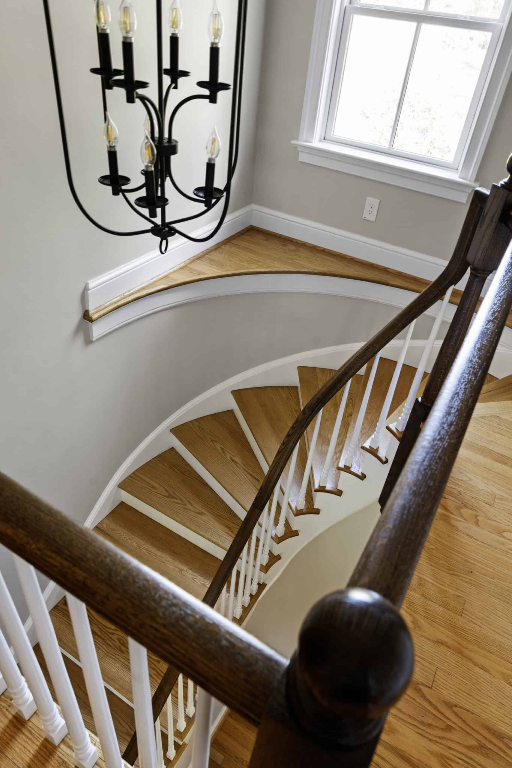 View from second floor of a white staircase with wood tread, white baluster, and wood guard rail. A black hanging light fixture and window are present.