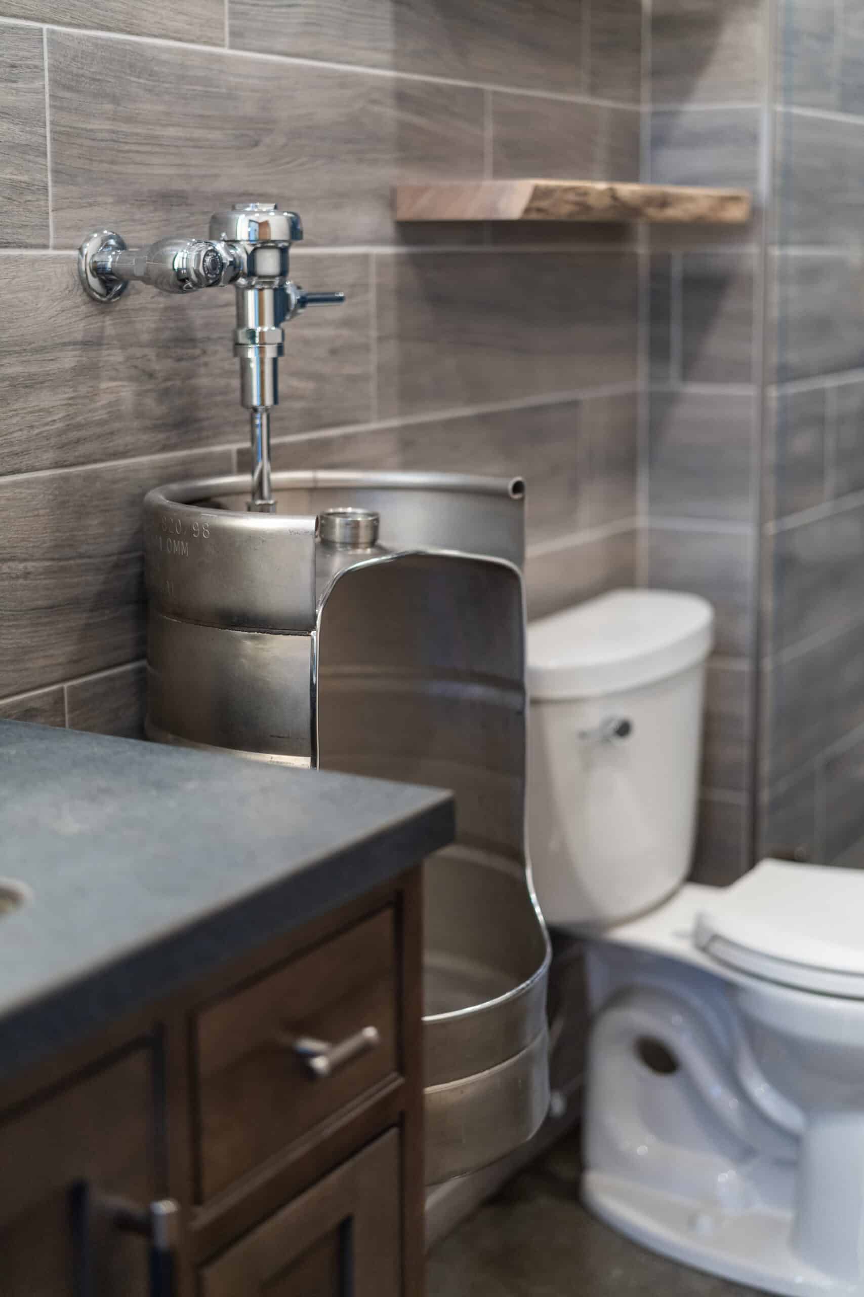 close-up shot of a unique urinal that appears to be  carved out of a metal keg. The bathroom has gray tile walls that look like brick work.