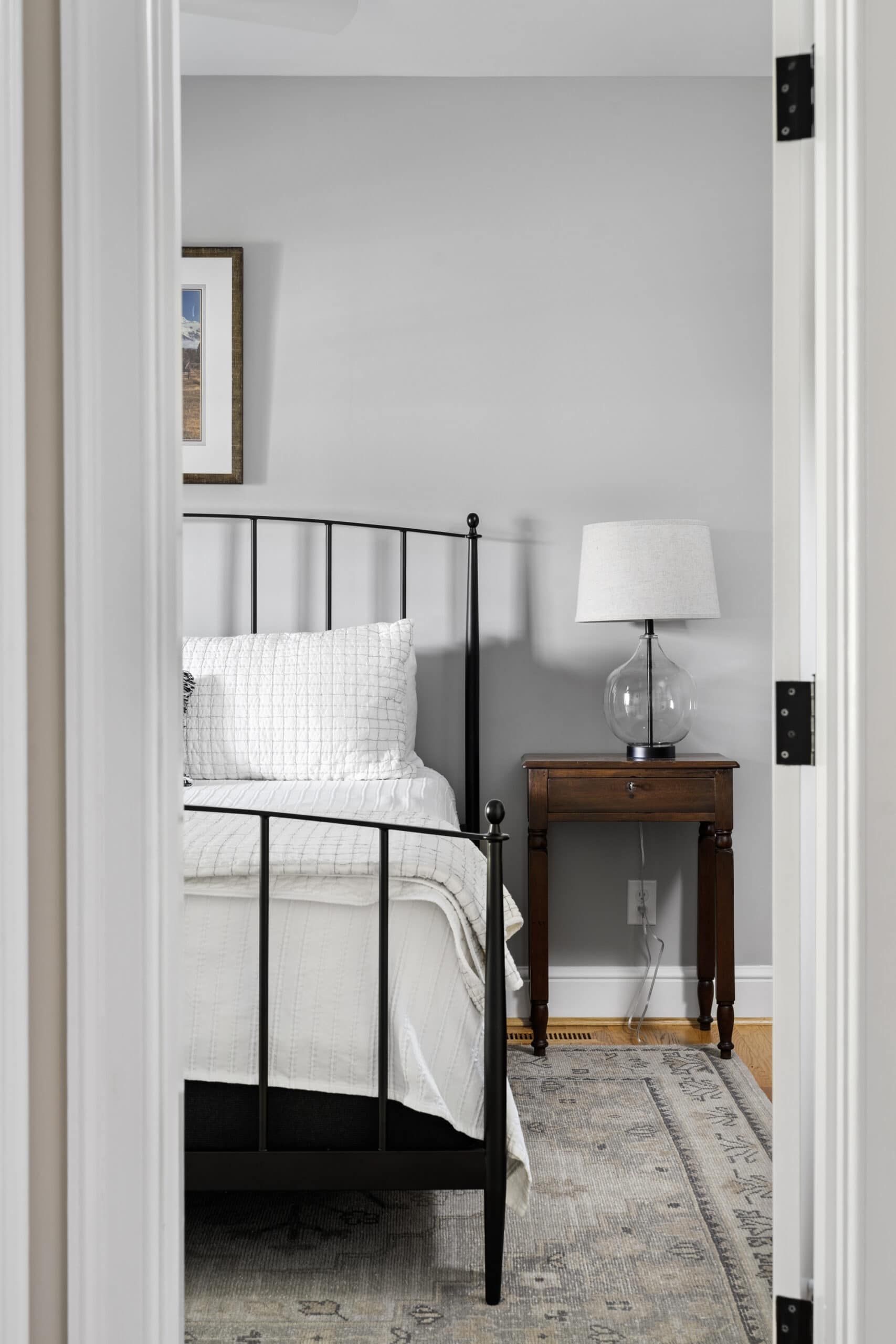 small bedroom seen through open door with black metal post bed frame, a small brown night stand, and a gray rug over wood floor