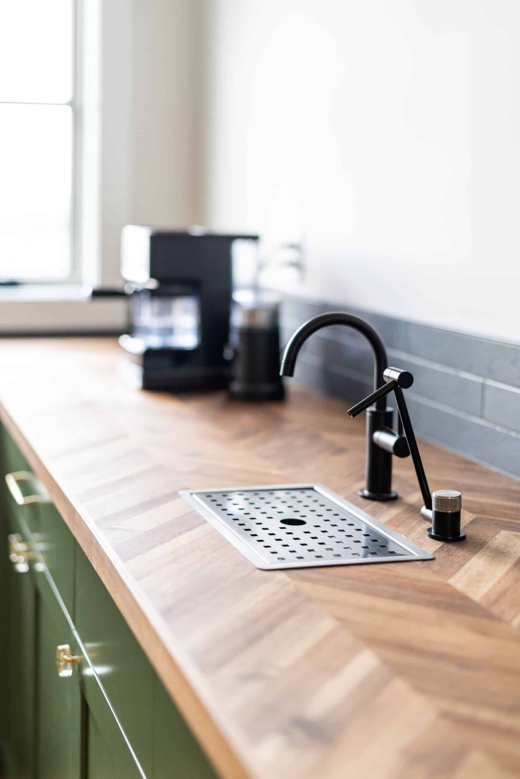 detail shot of a herringbone wood countertop with a black water faucet. A coffee maker and grinder are out of focus in the background.
