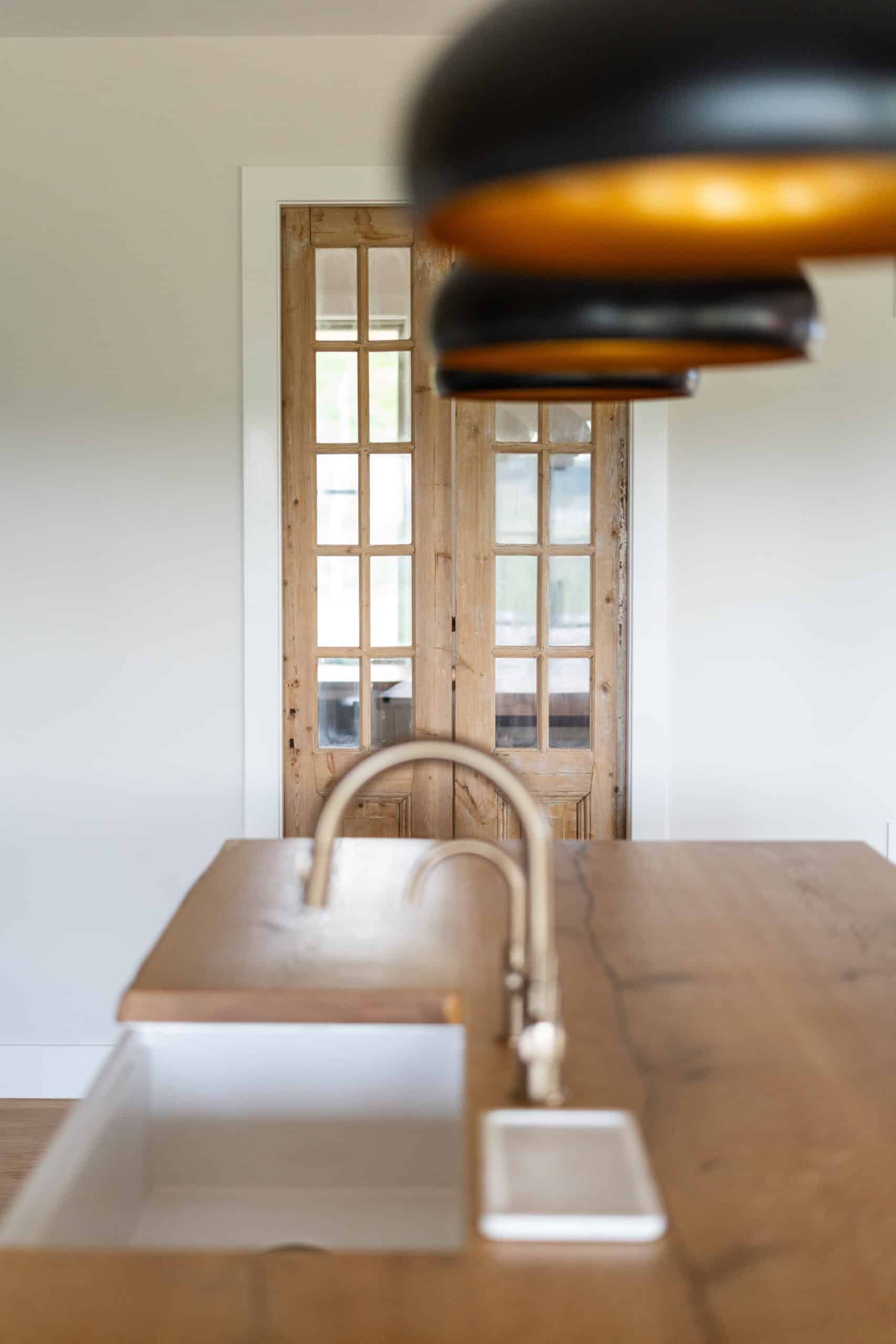 view of a wood and glass pantry door as seen from the opposite side of a kitchen island with a wood surface similar to the door. Three black overhead lights with orange light covers are out of focus