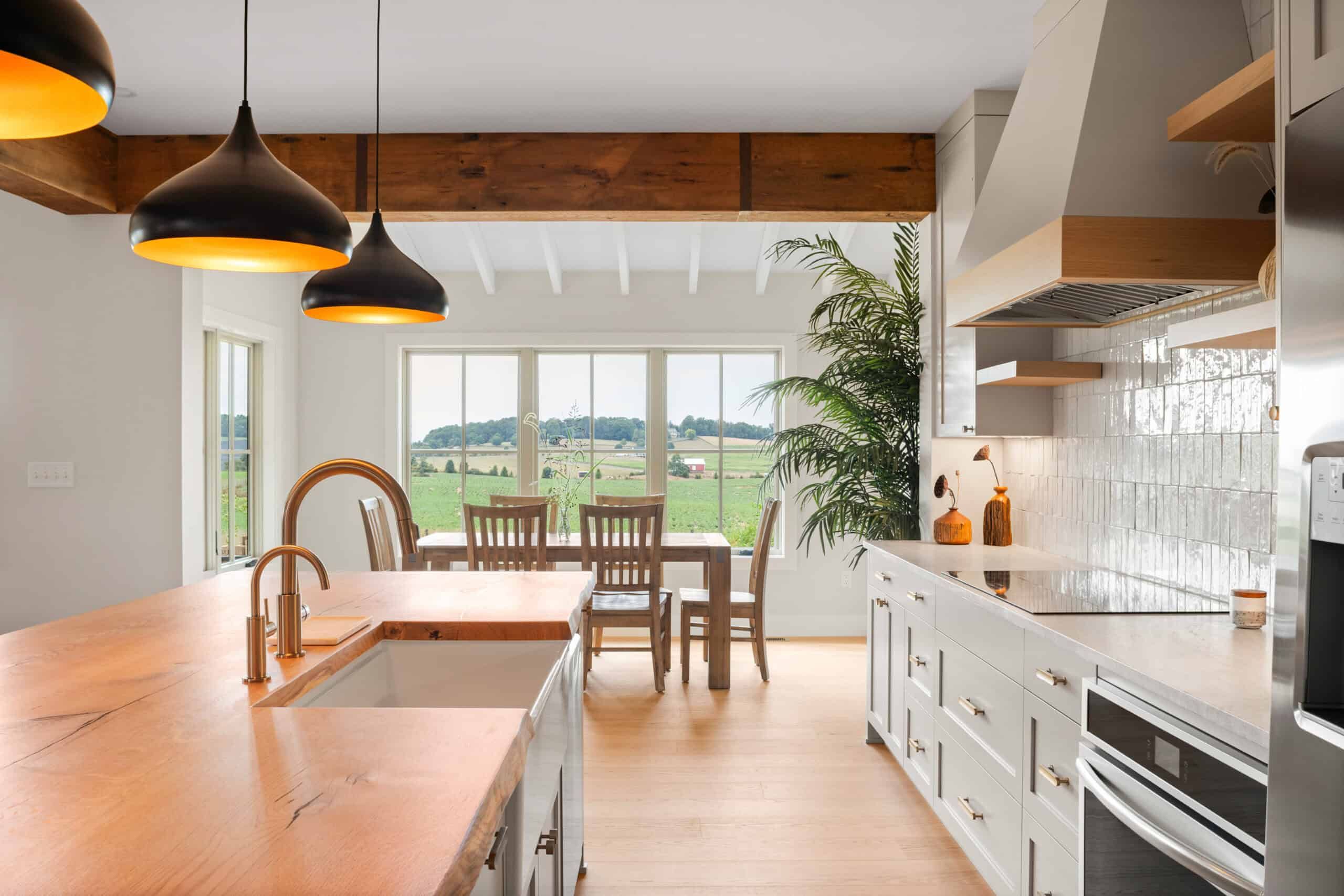 scenic green plains view out of of dining area window as seen from kitchen with off-white cabinets and a kitchen island. The room has brown wood floors matching the color of the kitchen island surface. Three black lamps with orange light covers are over the island.