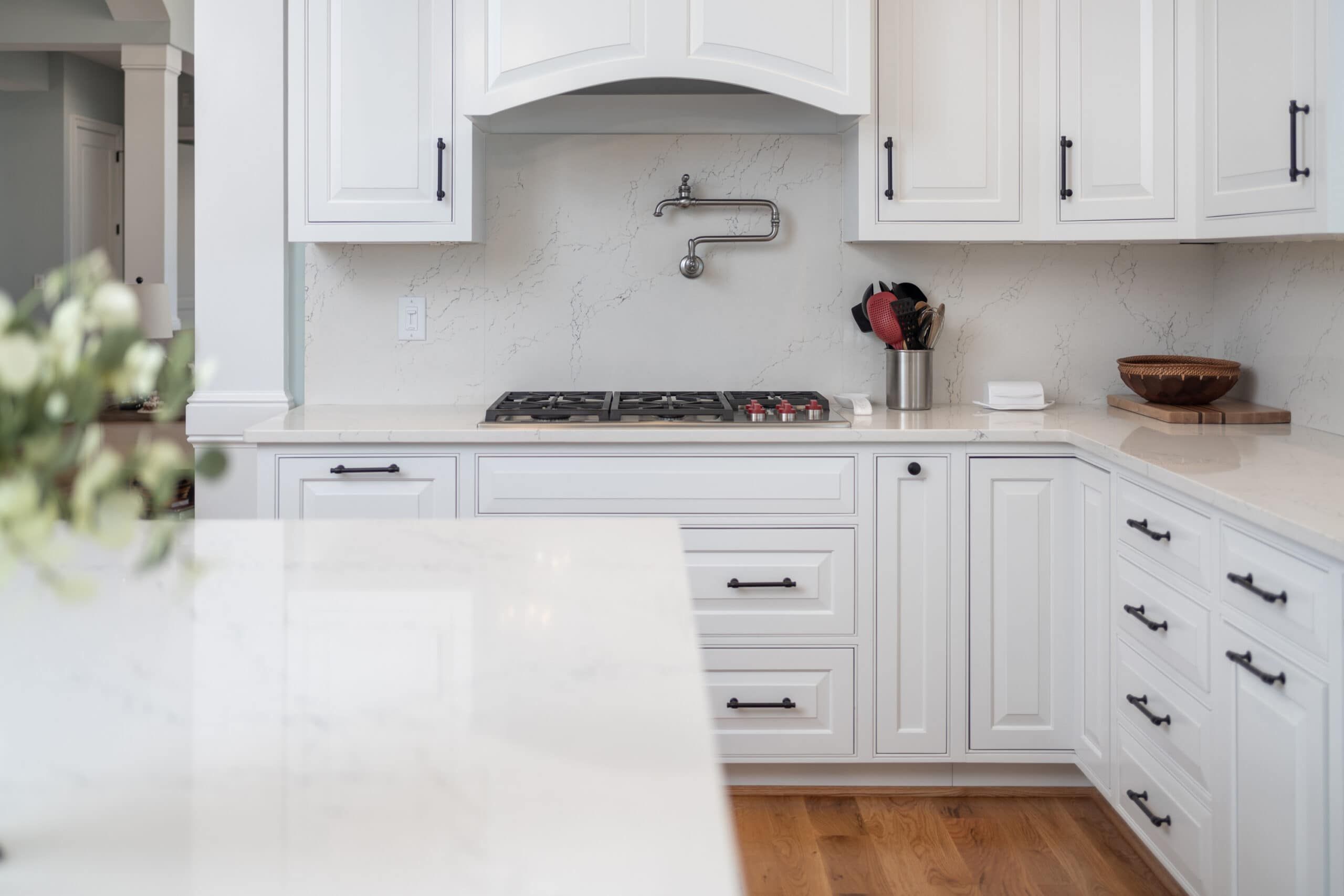 kitchen with a black and white motif, white quartz kitchen walls, white cabinets and black handles. A gas cooking surface is in focus, an interesting metal spigot with long pipe snaking at right angles is attached to the wall over the cooking surface. A shiny white kitchen island is blurred in the foreground, the floors are a medium brown wood
