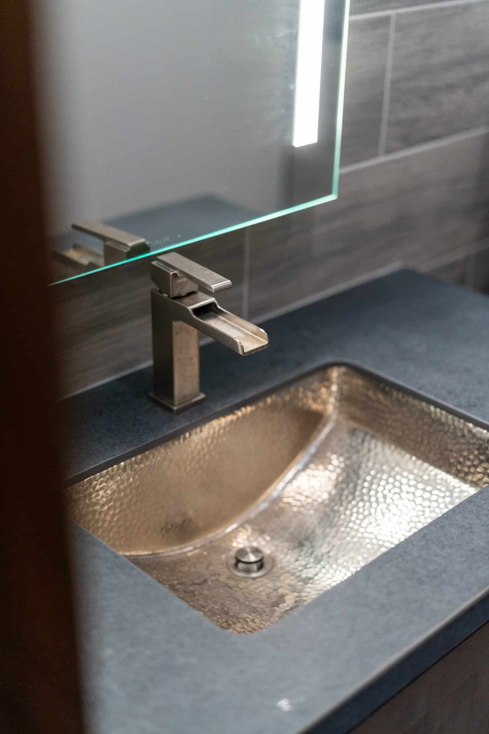 close-up of a metal bathroom sink basin, with square styled faucet beneath a mirror