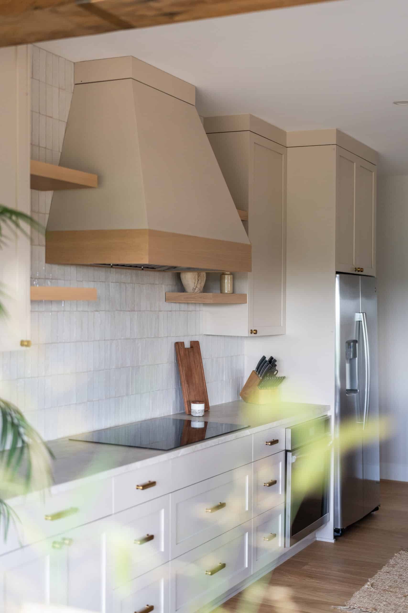 off-white kitchen cabinets and stove hood with light brown shelves attached to a shiny white, vertical tile wall. A glass smooth top cook surface and stainless steel appliances are present.