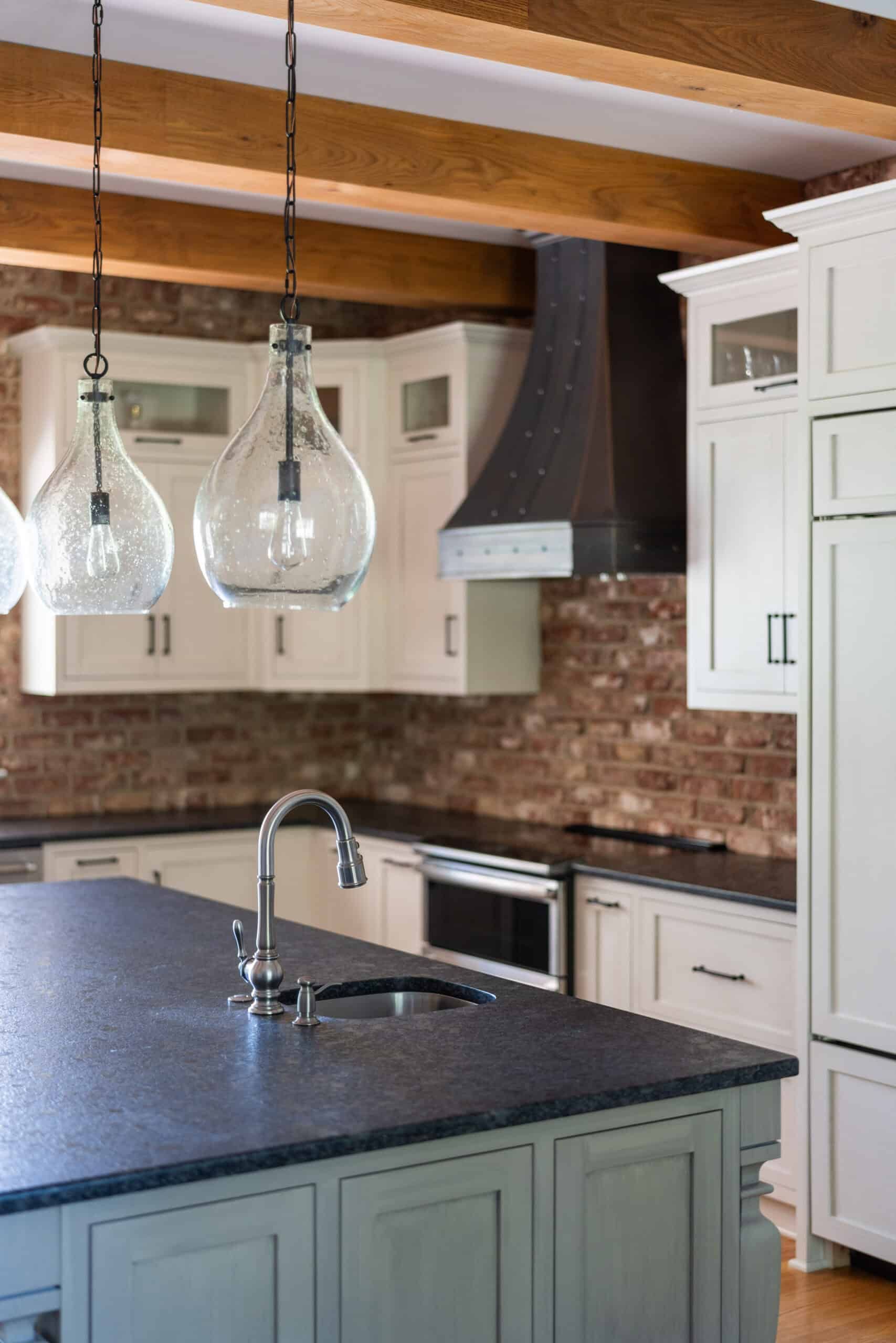 a natural brick kitchen with white cabinets, and a granite stove hood; a kitchen island with large hanging lights are in the foreground