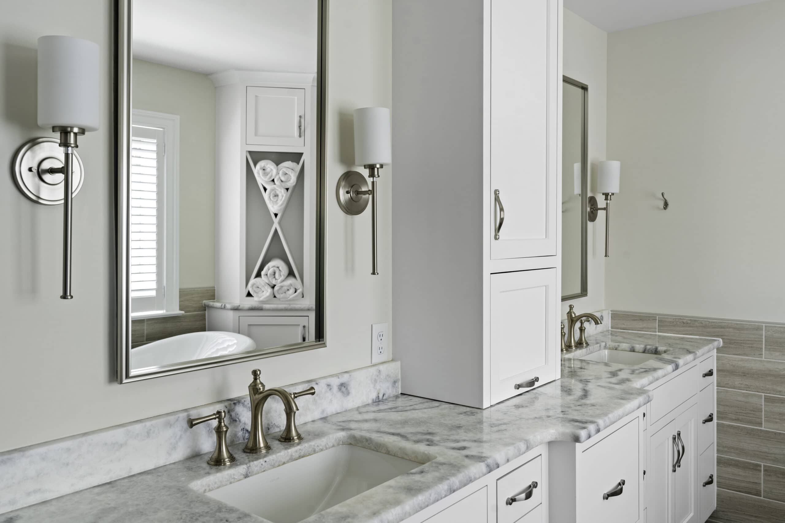 wide angle view of a bathroom with white granite countertops, white walls, white cabinets accented with metal handles, wall mounted lights, and mirror