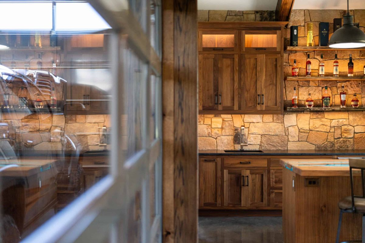 an in-home bar with natural stone walls and dark wood cabinets, to the left is a reflection of the room in a window