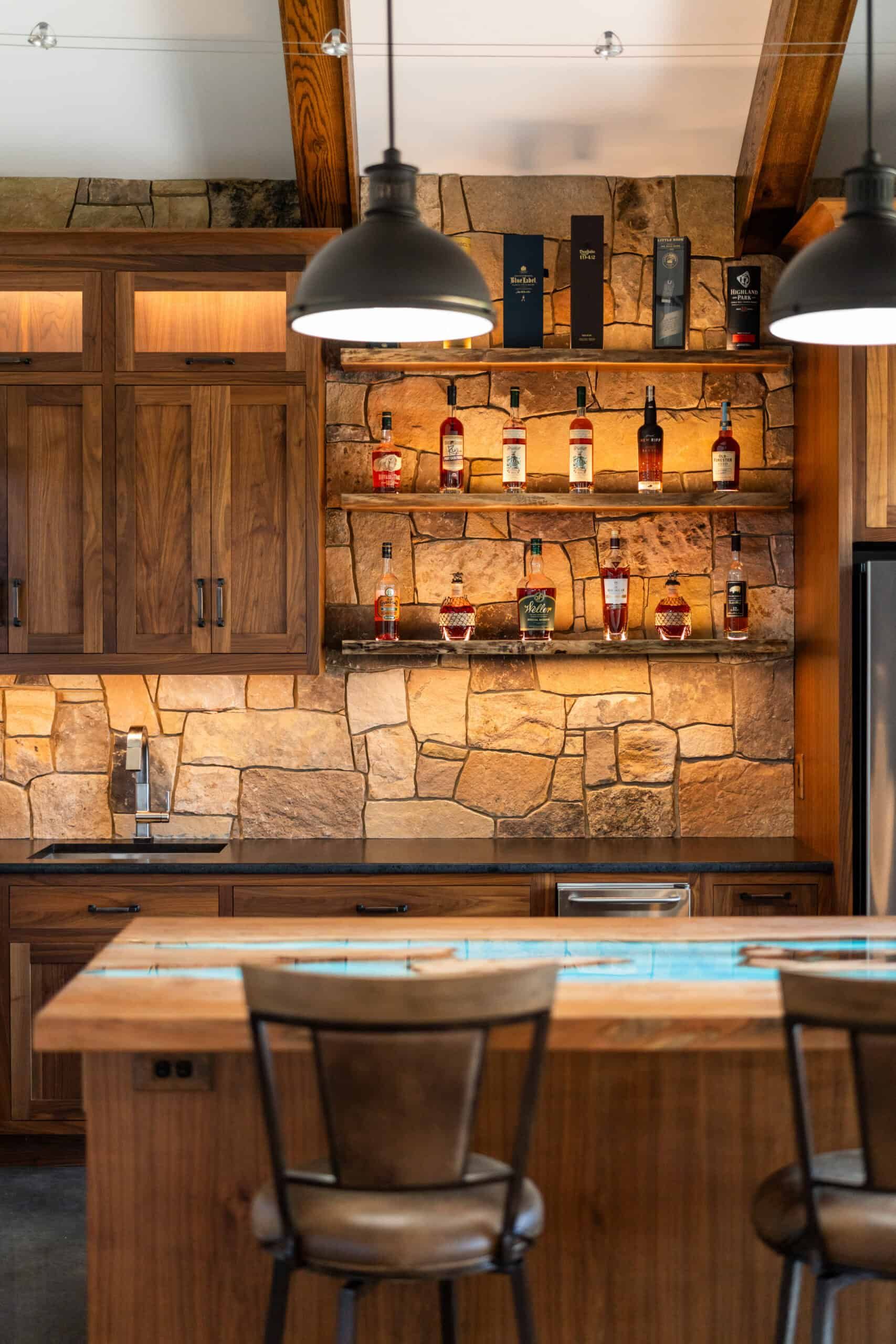 an in-home bar with natural stone walls and dark wood cabinets. Two stools are in front of a wood island that has a decorative effect evocative of bright, flowing water