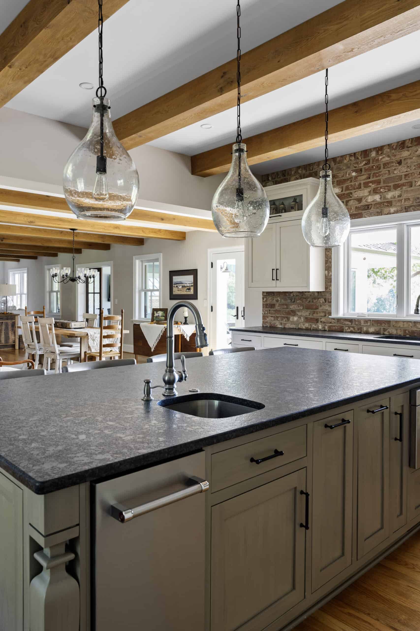 kitchen island with gray cabinets and black granite surface with three, transparent glass hanging lights in a natural brick kitchen with white cabinets, multiple thick, wood ceiling beams are prominent. The dining room leads to the visual horizon.
