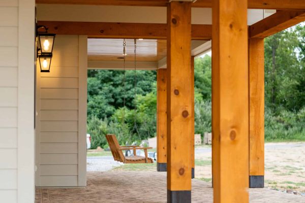 a swinging wooden bench on an outdoor patio, framed between a home's wall and four natural wood columns on a sunny day