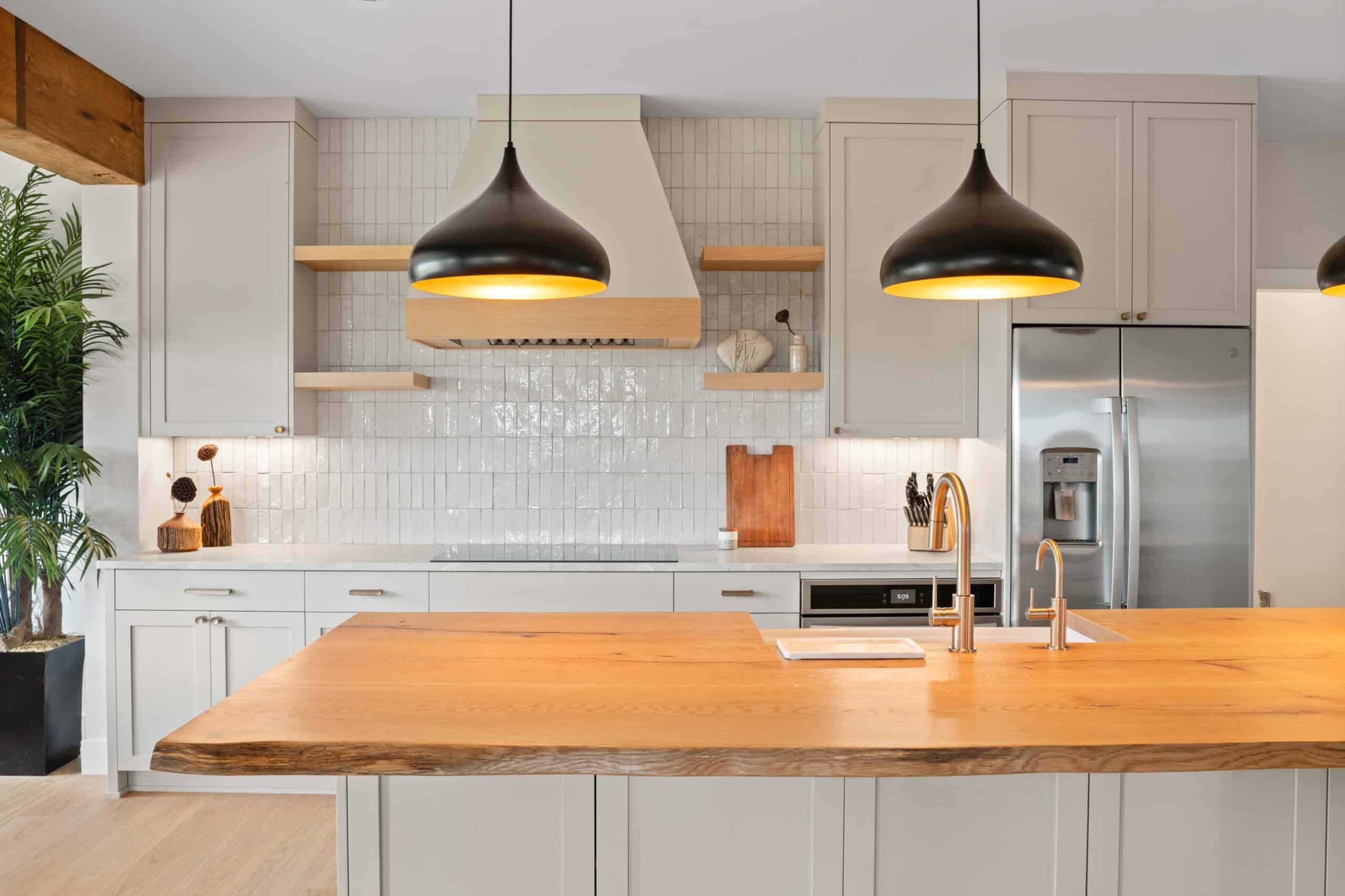 kitchen island with light brown wood surface and black hanging lamps with orange light covers. kitchen has shiny white, vertical tile walls, off-white cabinets, light wood shelves, and stainless steel appliances