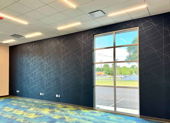 dark office wall with light contrasting stroke pattern and a floor to ceiling window. The carpet is a colorful geometric pattern, while the ceiling has thin bar lights some distance apart