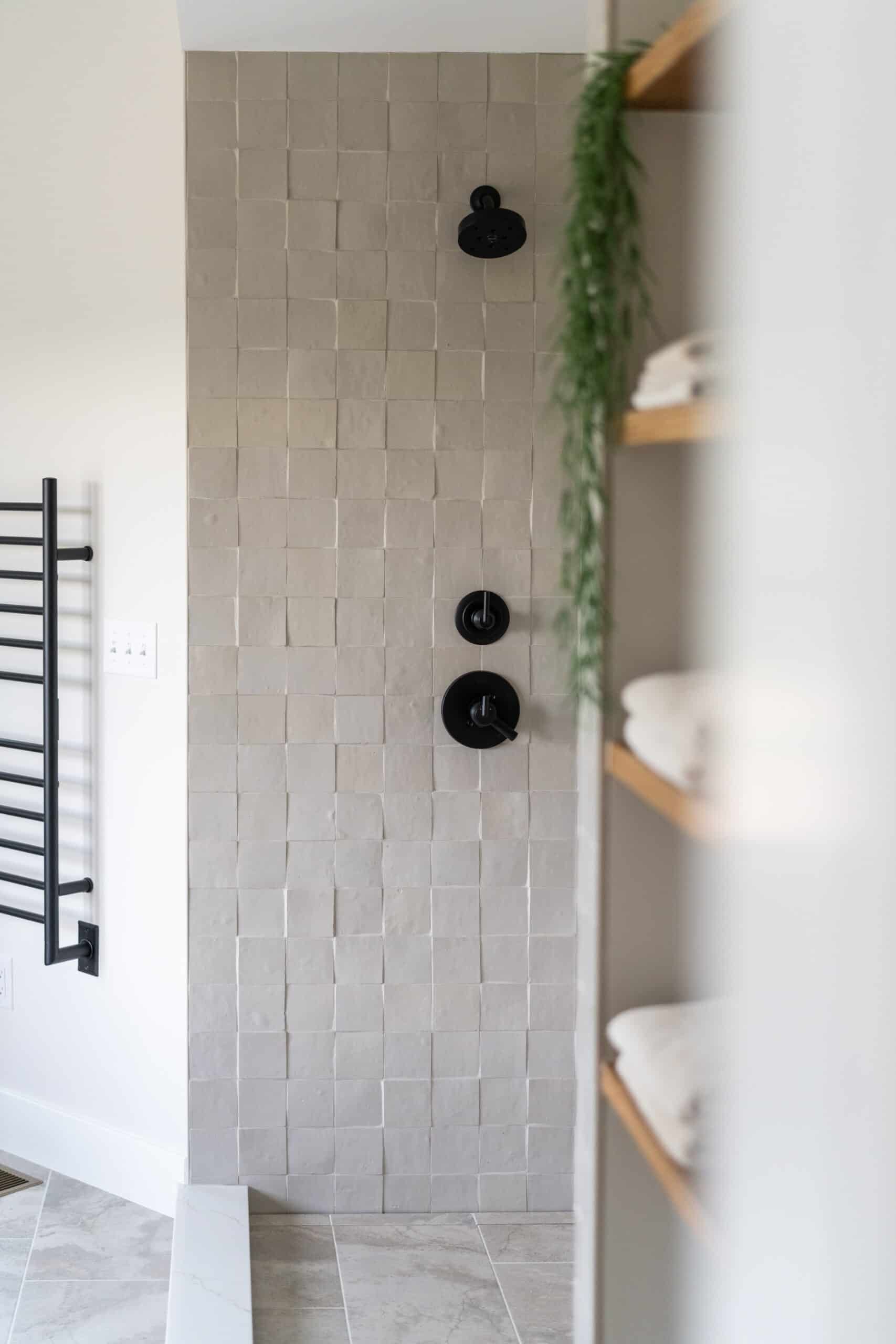 a wet room shower with white wall composed of square stones installed at alternating depths, the shower has black water features. a towel shelf with hanging green plant is slightly out of focus in the foreground 