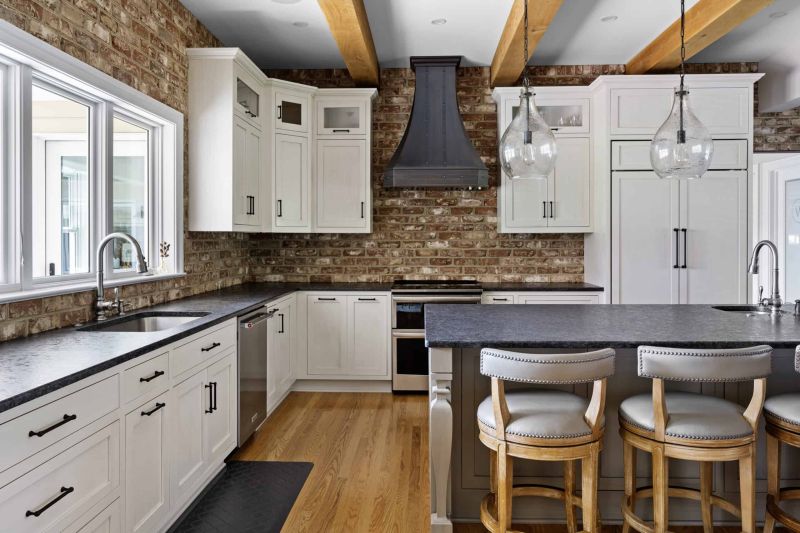 a natural brick kitchen with white cabinets, black countertops, and a kitchen island with two wood chairs and gray leather seats