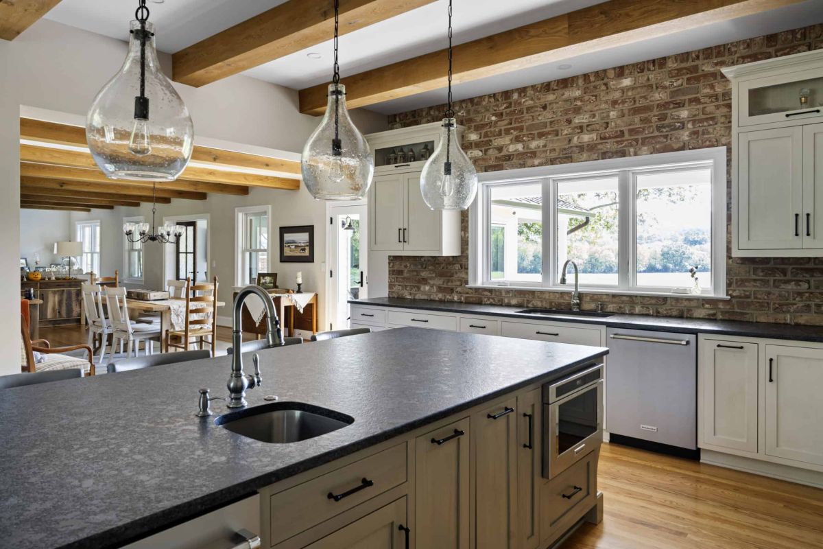 a kitchen and dining area lit by natural light from multiple windows, the kitchen has natural brick walls, white cabinets, and a gray kitchen island with a black granite surface. Over the island are three, transparent hanging lights. A dining room is on the visual horizon