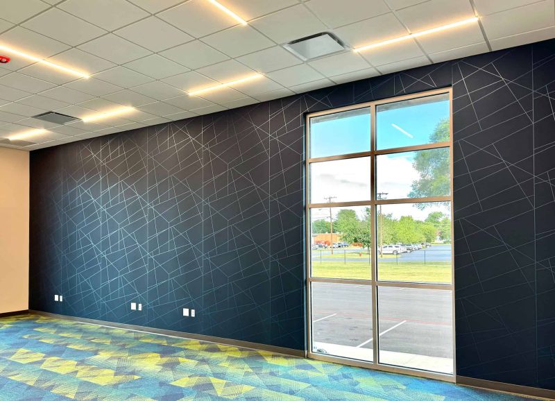 dark office wall with light contrasting stroke pattern and a floor to ceiling window. The carpet is a colorful geometric pattern, while the ceiling has thin bar lights some distance apart